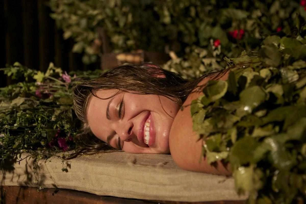 A smiling woman enjoying a soothing herbal body treatment with fresh sauna plants on a wooden bench.
